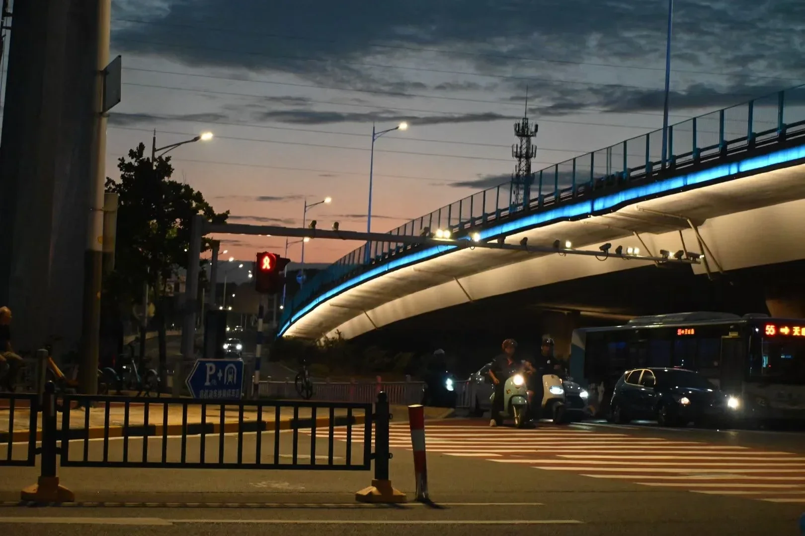 A person stands on a street beneath a bridge lit blue at dusk.